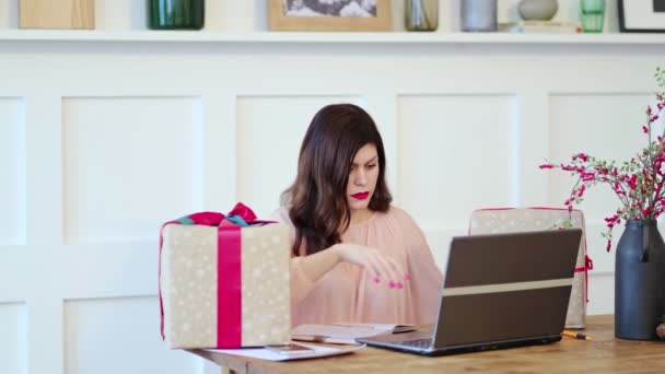 Masada oturan, dizüstü bilgisayarda yazan ve hediye kutularıyla konuşan güzel genç bir bayan. kutu evde iyi online alışveriş alır, posta sevkiyatı konsepti — Stok video