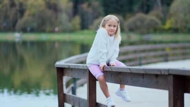 Portrait de jolie petite fille caucasienne aux cheveux blonds souriant à la caméra. Le gamin est assis sur une clôture. petite fille ne veut pas observer les stéréotypes imposés par la société — Video