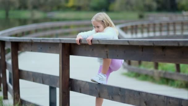 Portrait de jolie petite fille caucasienne aux cheveux blonds souriant à la caméra. Le gamin est assis sur une clôture. petite fille ne veut pas observer les stéréotypes imposés par la société — Video