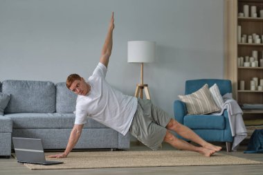 Beyaz tişörtlü kızıl adam oturma odasında çalışıyor. Paspasın üzerinde spor yapan bir adam oturma odasında dizüstü bilgisayarda videolar seyrediyor. Yerde su ve dambıllar var.