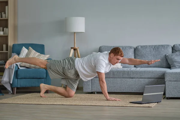 redhead man in white t-shirt works out in the living room. Man doing sports on a mat watching videos on a laptop in the living room with water and dumbbells on the floor