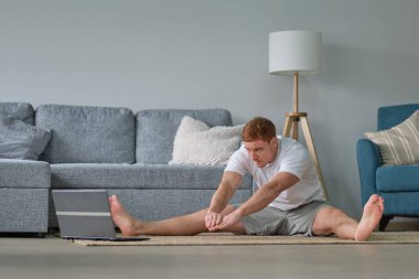 Beyaz tişörtlü kızıl adam oturma odasında çalışıyor. Paspasın üzerinde spor yapan bir adam oturma odasında dizüstü bilgisayarda videolar seyrediyor. Yerde su ve dambıllar var.