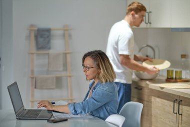 Modern çokuluslu çift mutfaklarında. Çalışan ya da çalışan kadınlar dizüstü bilgisayar kullanır. Arkadaki koca yemek pişiriyor ya da temizlik yapıyor. Cinsiyet kalıpları olmadan sağlıklı ilişkiler.