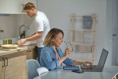 Modern çokuluslu çift mutfaklarında. Çalışan ya da çalışan kadınlar dizüstü bilgisayar kullanır. Arkadaki koca yemek pişiriyor ya da temizlik yapıyor. Cinsiyet kalıpları olmadan sağlıklı ilişkiler.