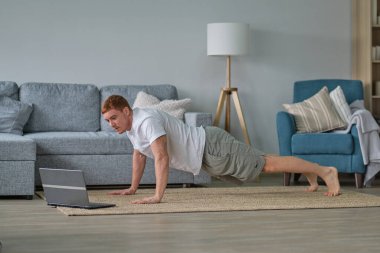 Beyaz tişörtlü kızıl adam oturma odasında çalışıyor. Paspasın üzerinde spor yapan bir adam oturma odasında dizüstü bilgisayarda videolar seyrediyor. Yerde su ve dambıllar var.