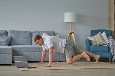 Beyaz tişörtlü kızıl adam oturma odasında çalışıyor. Paspasın üzerinde spor yapan bir adam oturma odasında dizüstü bilgisayarda videolar seyrediyor. Yerde su ve dambıllar var.