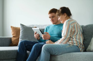 Oğul annesine tablet kullanmayı öğretiyor. Yaşlılar teknolojiyi kullanıyor. Kanepede, yetişkin oğlunun yanında oturan neşeli yaşlı kadın.