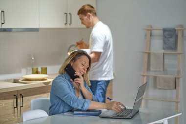 Meşgul iş kadını telefonda konuşuyor ve mutfaktaki bilgisayarı kullanıyor. Arkadaki ortak ya da koca yemek pişiriyor. sağlıklı ilişkiler modern çok kültürlü aile