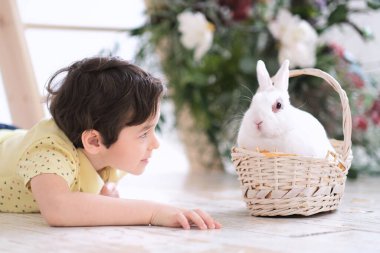 Küçük çocuk tahta zeminde uzanıyor ve beyaz bir tavşanla evcil bir hayvanla oynuyor. Tavşan hasır bir sepette oturur.