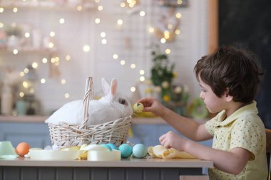 Mutlu Paskalyalar. Tavşan kulaklı çocuk süslü yumurtalarla oynuyor ve tavşan mutfak masasındaki hasır sepetin içinde oturuyor. yumuşak odak