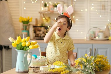 Paskalya yumurtası ve tavşan kulaklı neşeli çocuk mutfakta oynuyor. Mutlu çocuk Paskalya için hazırlanıyor.