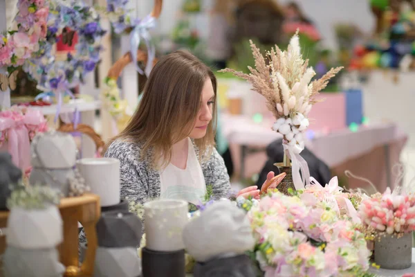 business owner selling behind counter with her bouquet of dried flowers at local market of craftsmen, small business. young woman entrepreneur sells floral holiday composition.