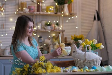 Genç bir kadın, Paskalya sepetini mutfak içinde dekore ediyor. Çiçekli mutfak, Paskalya yumurtaları, hediyeler, sepette oturan beyaz tavşan..