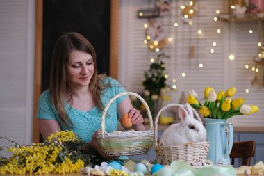 Genç bir kadın, Paskalya sepetini mutfak içinde dekore ediyor. Çiçekli mutfak, Paskalya yumurtaları, hediyeler, sepette oturan beyaz tavşan..