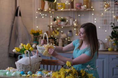 Genç bir kadın, Paskalya sepetini mutfak içinde dekore ediyor. Çiçekli mutfak, Paskalya yumurtaları, hediyeler, sepette oturan beyaz tavşan..