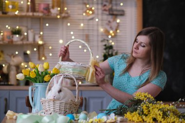 Genç bir kadın, Paskalya sepetini mutfak içinde dekore ediyor. Çiçekli mutfak, Paskalya yumurtaları, hediyeler, sepette oturan beyaz tavşan..