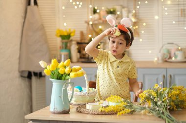 Tavşan kulaklı çocuk masada mutfakta oturuyordu. Paskalya için hazırlanan boyalı yumurtaları tutan çocuk. Başparmak gösteren komik çocuk.