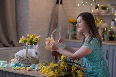 Genç bir kadın, Paskalya sepetini mutfak içinde dekore ediyor. Çiçekli mutfak, Paskalya yumurtaları, hediyeler, sepette oturan beyaz tavşan..