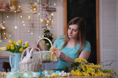 Genç bir kadın, Paskalya sepetini mutfak içinde dekore ediyor. Çiçekli mutfak, Paskalya yumurtaları, hediyeler, sepette oturan beyaz tavşan..