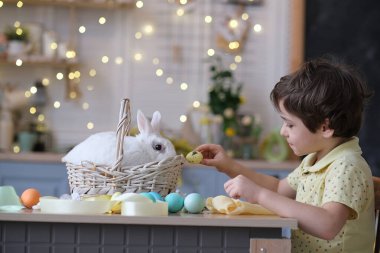 Mutlu Paskalyalar. Tavşan kulaklı çocuk süslü yumurtalarla oynuyor ve tavşan mutfak masasındaki hasır sepetin içinde oturuyor. yumuşak odak