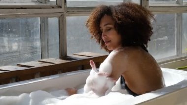 Gülümseyen ve rahatlatan Afro-Amerikan kadın köpük dolu bir küvette banyo yapıyor. Harika bir zaman. Yaşam tarzı insan kavramı