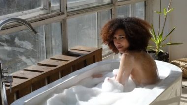 Gülümseyen ve rahatlatan Afro-Amerikan kadın köpük dolu bir küvette banyo yapıyor. Harika bir zaman. Yaşam tarzı insan kavramı