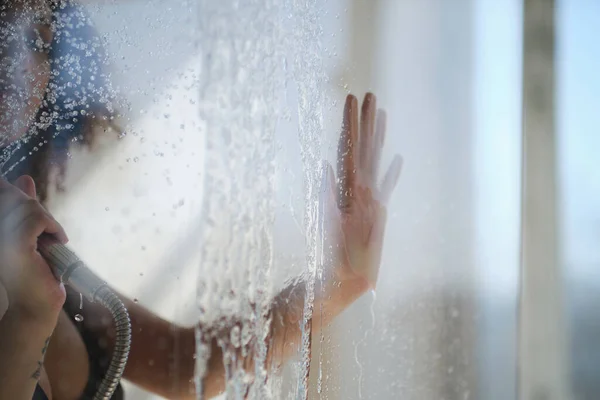 Női kéz a zuhanyfülke üvegajtaján. Érzéki portré fiatal nőről, amint zuhanyozik. Defokted női néz át az üvegen keresztül a zuhanyfülke. — Stock Fotó