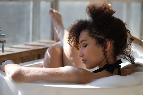 Vista trasera chica negra rizado por la que se extiende, relajarse en el baño. Cuidado de la belleza, tratamientos de spa y relajarse en casa. Alegre joven afroamericana mujer disfrutando de agua caliente en el interior del baño por la mañana. — Foto de Stock