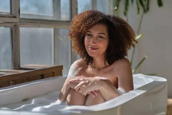 Mosolygó és pihentető afro-amerikai nő fürdik egy kádban tele habbal. Csodálatos volt. életmód emberek koncepció — Stock Fotó