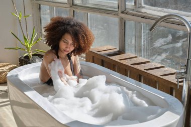 Gülümseyen ve rahatlatan Afro-Amerikan kadın köpük dolu bir küvette banyo yapıyor. Harika bir zaman. Yaşam tarzı insan kavramı