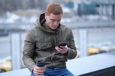 Genç adam akıllı telefon kullanıyor ve bluetooth kulaklıkla müzik dinliyor. Sonbahar ceketli yalnız adam şehirde yürürken kahve içer.