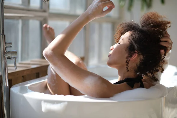 Vista trasera chica negra rizado por la que se extiende, relajarse en el baño en el interior. mujer joven que se relaja en bañera caliente con espuma y burbujas. Mujer cansada disfrutando del descanso mimarse. — Foto de Stock