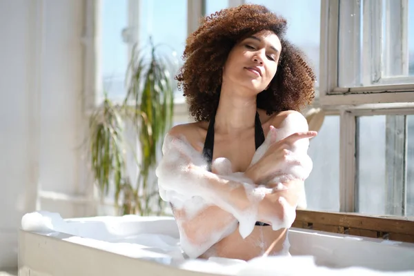 Gülümseyen ve rahatlatan Afro-Amerikan kadın köpük dolu bir küvette banyo yapıyor. Harika bir zaman. Yaşam tarzı insan kavramı