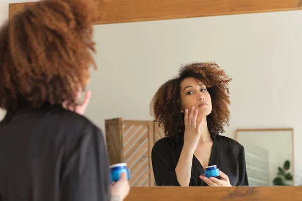 Junge Frau Anwendung Kosmetik Badezimmer. Weibliche Gesichter im Spiegel. Schöne Afroamerikanerin trägt Hautpflegecreme auf das Gesicht auf. — Stockfoto