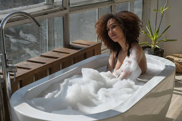 Donna afroamericana sorridente e rilassante che fa il bagno in una vasca piena di schiuma. Che bella serata. stile di vita persone concetto — Foto Stock