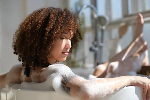 Vue arrière fille noire bouclée pose, détente dans la salle de bain à l'intérieur. jeune femme relaxante dans une baignoire chaude avec mousse et bulles. Fatigué femelle profiter repos choyer elle-même. — Photo