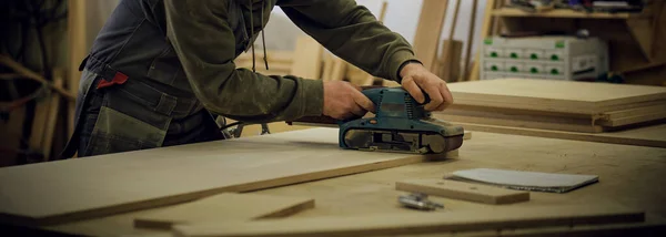 De cerca. Carpintero en taller pule tablero de madera con una lijadora orbital eléctrica. Concepto de carpintería y mobiliario. — Foto de Stock