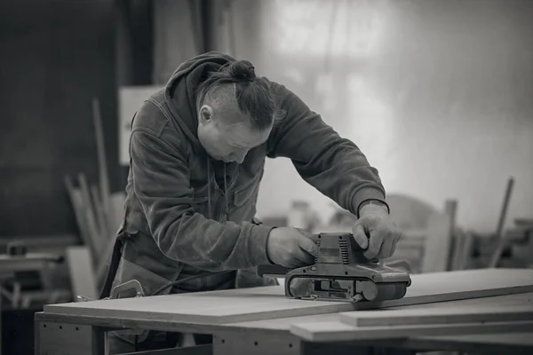 De cerca. Carpintero en taller pule tablero de madera con una lijadora orbital eléctrica. Concepto de carpintería y fabricación de muebles. fotografía en blanco y negro — Foto de Stock