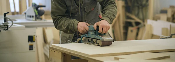 De cerca. Carpintero en taller pule tablero de madera con una lijadora orbital eléctrica. Concepto de carpintería y mobiliario. — Foto de Stock