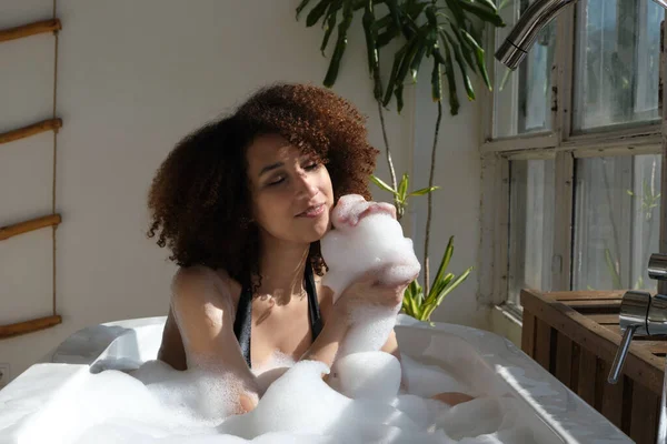微笑和放松的非洲裔美国女人沐浴在充满泡沫的浴缸里。令人惊奇的时刻生活方式概念 — 图库照片