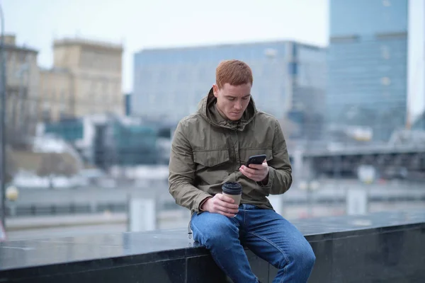 Hombre joven usando el teléfono inteligente y escucha música en los auriculares del bluetooth. hombre solitario en chaqueta de otoño bebe café mientras camina por la ciudad — Foto de Stock