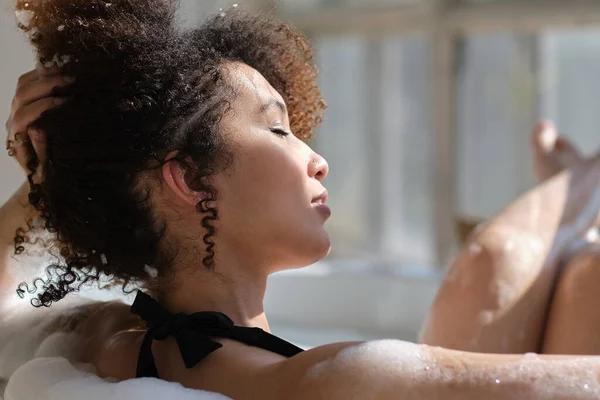 Visão traseira encaracolado menina negra deitado, relaxante no banho dentro de casa. jovem relaxante na banheira quente com espuma e bolhas. Cansada fêmea desfrutando de descanso mimar-se. — Fotografia de Stock