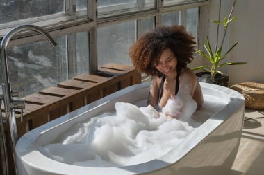 Gülümseyen ve rahatlatan Afro-Amerikan kadın köpük dolu bir küvette banyo yapıyor. Harika bir zaman. Yaşam tarzı insan kavramı