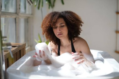 Gülümseyen ve rahatlatan Afro-Amerikan kadın köpük dolu bir küvette banyo yapıyor. Harika bir zaman. Yaşam tarzı insan kavramı