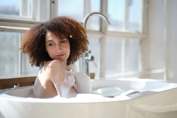 Χαμογελώντας και χαλαρώνοντας Αφροαμερικανή γυναίκα που κάνει μπάνιο σε μια μπανιέρα γεμάτη αφρό. Καταπληκτικές στιγμές. lifestyle άνθρωποι έννοια — Φωτογραφία Αρχείου