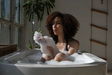 Gülümseyen ve rahatlatan Afro-Amerikan kadın köpük dolu bir küvette banyo yapıyor. Harika bir zaman. Yaşam tarzı insan kavramı