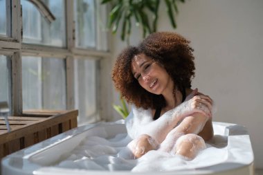 Gülümseyen ve rahatlatan Afro-Amerikan kadın köpük dolu bir küvette banyo yapıyor. Harika bir zaman. Yaşam tarzı insan kavramı