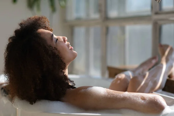 Visão traseira encaracolado menina negra deitado, relaxante no banho. Cuidados de beleza, tratamentos de spa e relaxar em casa. Jovem mulher afro-americana alegre desfrutando de água morna no interior do banheiro de manhã. — Fotografia de Stock