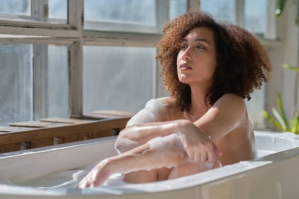 Femme afro-américaine souriante et relaxante se baignant dans une baignoire pleine de mousse. Un temps incroyable. style de vie gens concept — Photo
