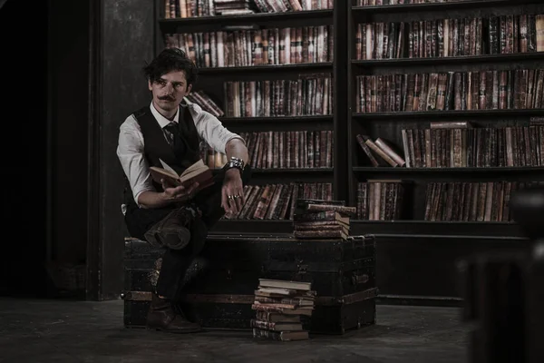 pensive man stands at night in a dark library room and holding a vintage book. stylish man with a mustache straightens the bracelet.